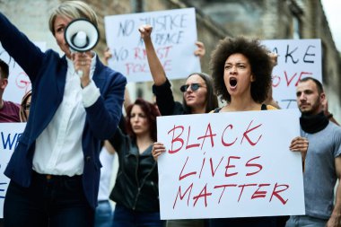 Large group of multi-ethnic people demonstrating against racism through city streets. Focus is on black woman shouting while holding a banner. 