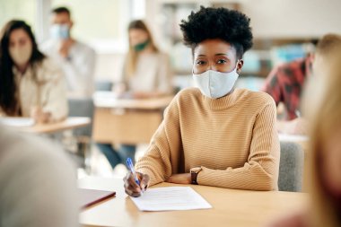 African American female student having an exam at the university and wearing face mask due to coronavirus pandemic.