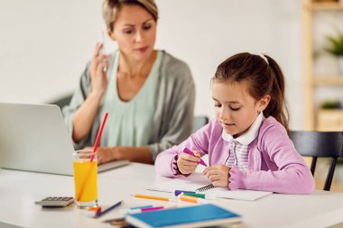 Cute small girl doing homework and drawing while mother is working on laptop next to her at home. 