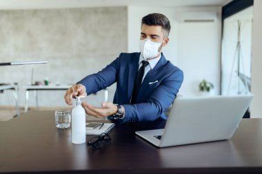 Businessman disinfecting his hands and wearing protective mask on his face while working in the office during coronavirus epidemic. 