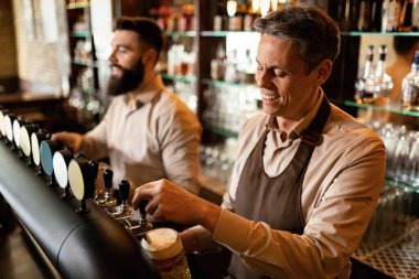 Happy barista pouring draft beer in a glass while working with a colleague in a bar. 