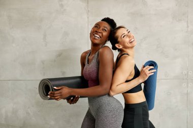 Happy sportswomen laughing after training class while standing against the wall and holding exercise mats. Copy space. 