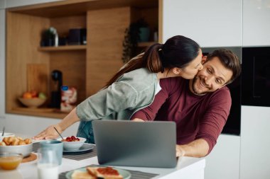 Happy couple having fun during breakfast at home. Woman is kissing her husband while he is surfing the net on laptop. 