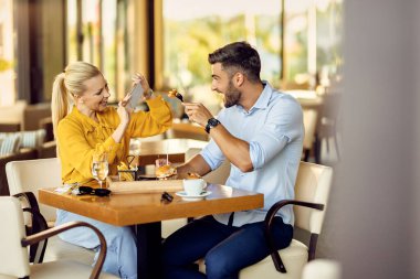 Cheerful man and fun with his girlfriend while she is using smart phone and taking picture of him during a lunch.