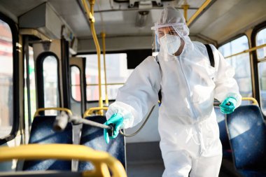 Healthcare worker in protective suit doing disinfection of public bus during coronavirus pandemic.