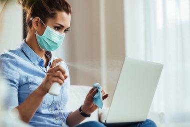 Young woman wearing protective face mask and using spray bottle while cleaning her laptop during virus epidemic. 