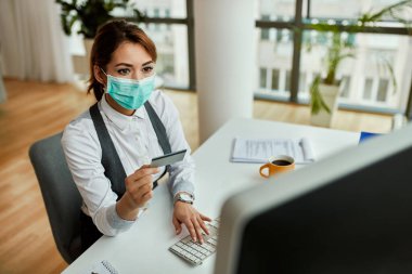 Young businesswoman with face mask using credit card and computer while e-banking in the office. 