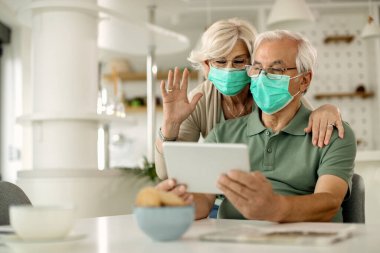 Senior couple having video call over digital tablet while wearing protective face masks due to coronavirus epidemic. 