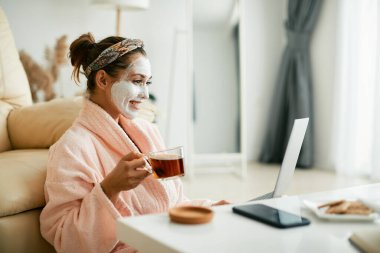 Bornozlu genç bir kadın, kozmetik maske takıp evde çay içerken dizüstü bilgisayarında internette sörf yapıyor..