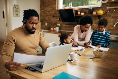Black father using laptop and working on paperwork while his wife is assisting their children in learning at home.