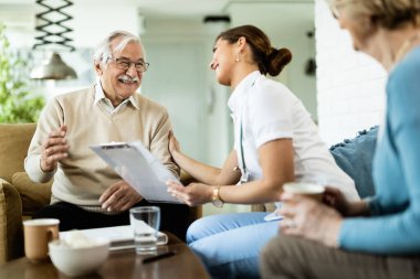 Happy senior man and young female doctor talking during home visit. 