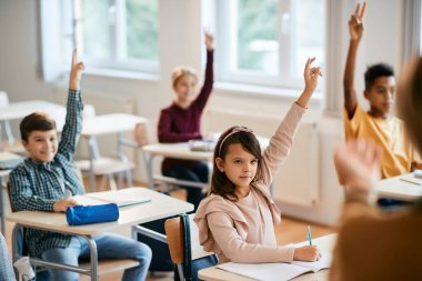 Schoolgirl and her classmates raising their arms to answer a question during a class in the classroom.