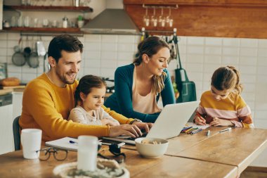 Happy parents spending time with their kids at home. One of daughters and father are using laptop while mother and other daughter are coloring on the paper. 