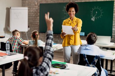 Happy African American teacher holding a class and pointing at student with raised arm who wants to answer the question. 
