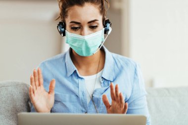 Young woman wearing protective face mask while using computer and having video call at home. 
