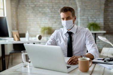 Male entrepreneur with protective face mask using computer while working at his office desk. 
