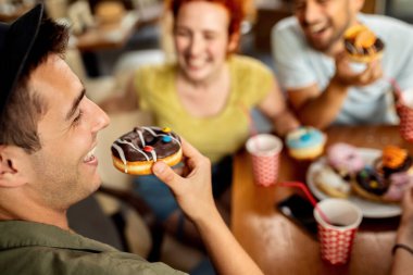 Young man having fun while eating donut and talking to his friends in a cafe. 
