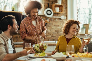 Group of young happy friends enjoying in lunch. Focus is on African American woman serving salad at dining table. 