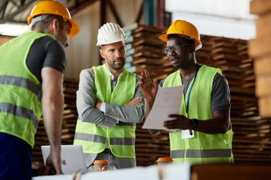 African American worker and his colleague talking to warehouse inspector while going through distribution plans at timber department.