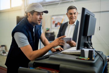 Auto repairman working on computer while talking with his manager in a workshop. 