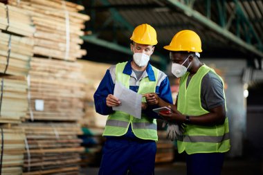 Two manual workers with face masks going though checklist while working in timber warehouse. 