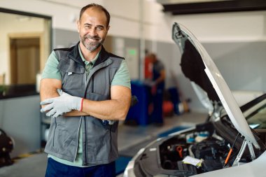 Happy auto mechanic standing with arms crossed and looking at camera while working in repair shop. 