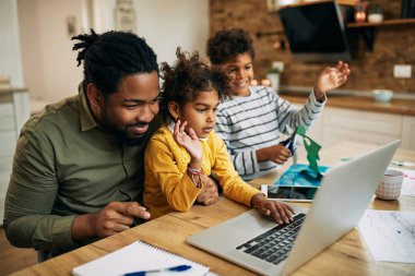 Happy African American father and children waving while greeting someone during video call over laptop. 