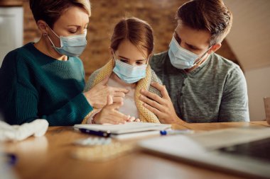 Worried parents taking care of their ill daughter at home and wearing protective face masks due to COVID-19 pandemic. 