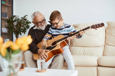 Happy little boy enjoying while his grandfather is teaching him to play acoustic guitar at home. 