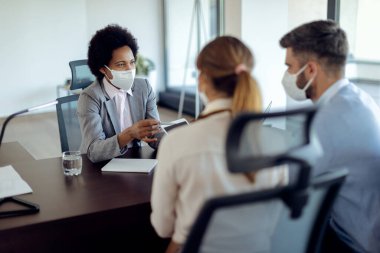 African American real estate agent using digital tablet while talking to a couple on a meeting in the office. They are wearing protective face masks due to COVID-19 epidemic. 