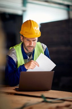 Mid adult foreman working on laptop while analyzing paperwork at distribution warehouse.