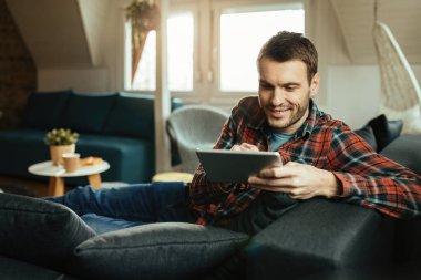 Evdeki kanepede dinlenirken dijital tablet kullanan gülümseyen genç adam..