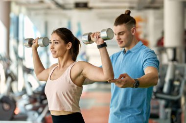 Spor salonundaki spor eğitiminde koçu ona yardımcı olurken genç sporcu el ağırlığı kullanıyor..