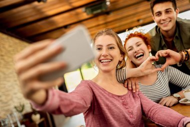 Low angle view of happy friends having fun and taking selfie with mobile phone in a cafe, 