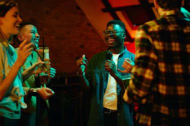 Group of happy friends having fun in a bar at night. Focus is on African American man signing karaoke. 