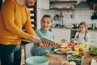 Happy husband showing something on touchpad to his while while she is preparing meal at dining table. Focus is on woman.