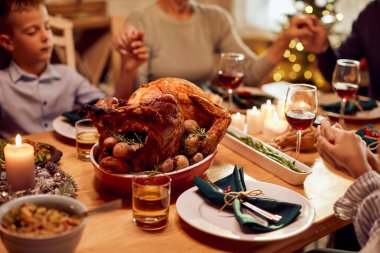Close-up of traditional roast turkey with family holding hands and praying during Thanksgiving dinner at dining table.