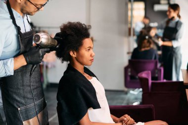 Male hairdresser styling African American woman's hair during appointment at the salon.