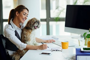 Young happy businesswoman working in the office while dog is sitting in her lap. 