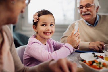Happy little girl communicating with her grandparents while using cell phone during lunch at dining table. 
