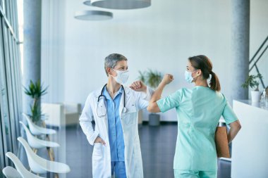 Mutlu kadın doktor ve hemşire, Coronavirus salgını sırasında klinikte dirsek ve yüz maskeleri takarak selamlıyorlar.. 