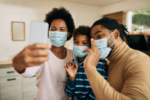 Happy black parents and their son having fun while taking selfie with mobile phone and wearing face masks due to coronavirus pandemic.
