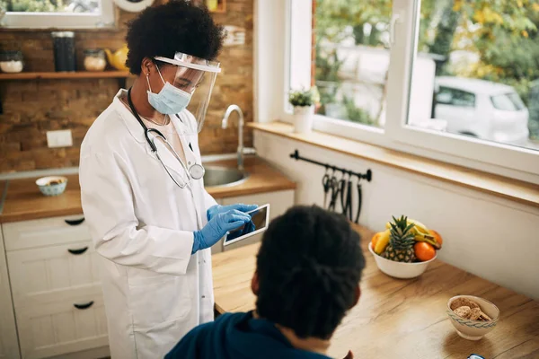 Black female doctor wearing face shield and mask while using digital tablet during a home visit.