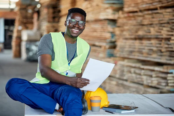 Happy African American warehouse worker analyzing distribution plans at lumber department and looking at camera.
