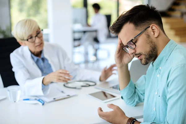 Distraught man holding his head in pain while talking to doctor at the clinic.