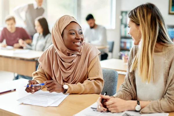 Happy female university friends communicating during a class in the classroom. Focus is on African American Islamic student.