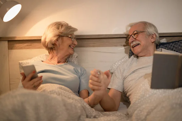 Happy senior couple talking to each other while holding hands and resting in the bedroom at night.