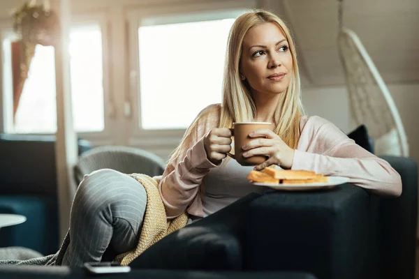 Smiling pensive woman relaxing on the sofa and enjoying in cup of coffee in the living room.