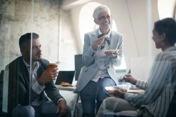 Happy Mature Businesswoman Eating Donut Communicating Her Coworkers Break Office — Zdjęcie stockowe