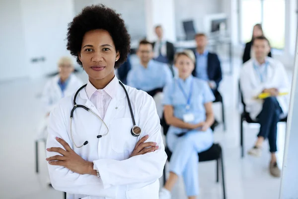 Smiling black doctor standing with her arms crossed while leading educational event in convention center and looking at camera.